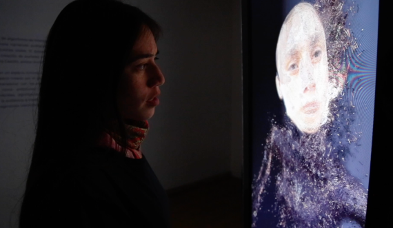 Image of Anaid Bohor, performer, controlling a virtual Avatar. Standing in front of a screen. 