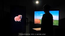 A person stands in silhouette between two large monitors in a dark gallery space. The left screen displays an animated guinea pig with a human face embedded into its head. The right screen shows a bright virtual field under a blue sky. Subtitles at the bottom read “[transforming into a guinea pig],” indicating the narrative or interactive transformation taking place.
