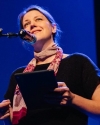 image of white woman wearing black shirt and scarf, holding a tablet and standing before a microphone