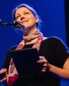 image of white woman wearing black shirt and scarf, holding a tablet and standing before a microphone