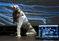 Real-time human-machine interaction during rehearsal. A dance artist performs in front of a projection controlled by our system described in the paper. The laptop screen shows the multimedia interface, while wearable IMU sensors and a smartphone are used to stream and monitor real-time data.